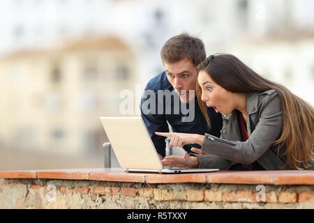 Surprised couple checking laptop content in a balcony with a town in the background Stock Photo