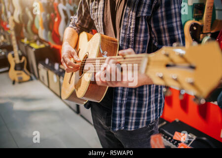 Close up of man standing and playing on acoustic guitar. He dresses up like hipster. Guy is alone in room Stock Photo