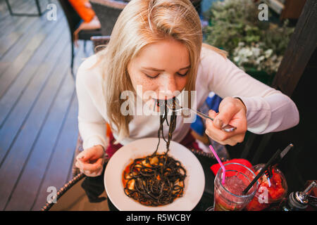 Young beautiful woman eating black pasta with seafood and cuttlefish ink in the outdoor restaurant. Funny and beautiful. Stock Photo