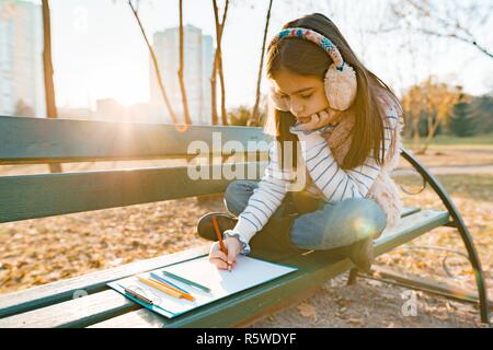 Little beautiful artist drawing with colored pencils, girl sitting on a bench in sunny autumn park, golden hour, Stock Photo