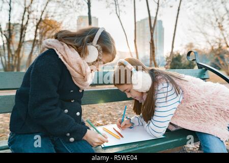 Two little pretty artists drawing with colored pencils, girls sitting on a bench in sunny autumn park, golden hour. Stock Photo