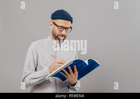 Serious male teacher writes down notes for next class, holds notepad, prepares interesting task for students, wears formal outfit, stands against grey Stock Photo