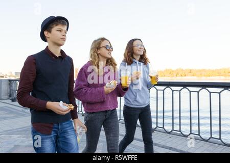 Lifestyle of adolescents, boy and two teen girls are walking in the city. Laughing, talking children eating street food, having fun. Background of the Stock Photo