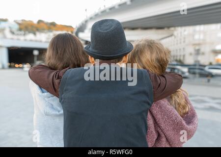 Three friends, view from the back of young teenager boy hugging two girls by the shoulders. Children look ahead, city life