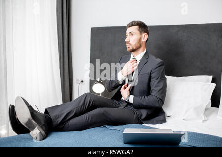 Elegant businessman in the suit fixing tie sitting during the work on the bed of the hotel room Stock Photo