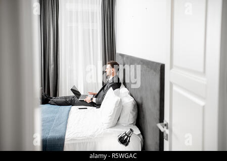 Elegant businessman in the suit working with laptop on the bed of the hotel room during a business trip. Wide view with copy space Stock Photo