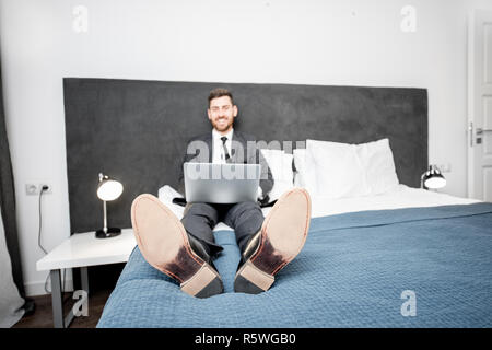 Funny portrait of a businessman in the suit working with laptop on the bed of the hotel room during a business trip. Image focused on the shoes Stock Photo