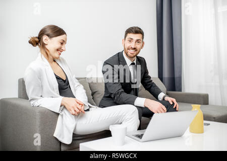 Elegant man and woman dressed in the suits doing business sitting together on the couch with laptop at home or comfortable office Stock Photo