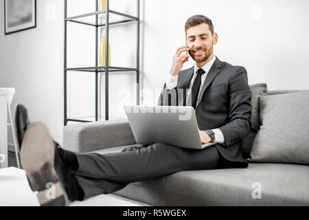 Elegant businessman dressed in the suit working with laptop and talking phone on the couch at home or comfortable office Stock Photo