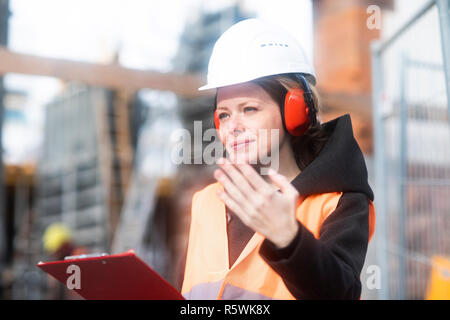 Portrait of a woman in a hard hat holding a clipboard and gesturing Stock Photo
