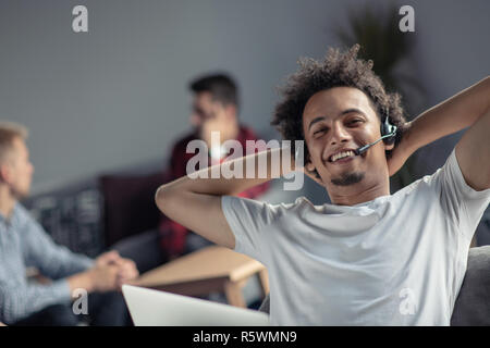Satisfied relaxed african-american freelancer feeling happy at work sitting at office desk with headset. Stock Photo