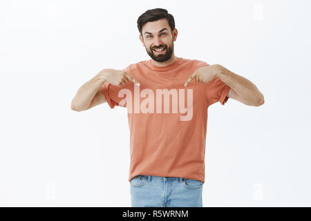 Portrait of satisfied good-looking carefree dark-haired bearded male in 30s, pointing down or t-shirt with happy and joyful smile gazing at camera sho Stock Photo