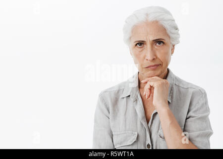 Waist-up shot of concerned serious-looking perplexed elderly woman with grey hair frowning and smirking from dissatisfaction holding hand on chin thin Stock Photo