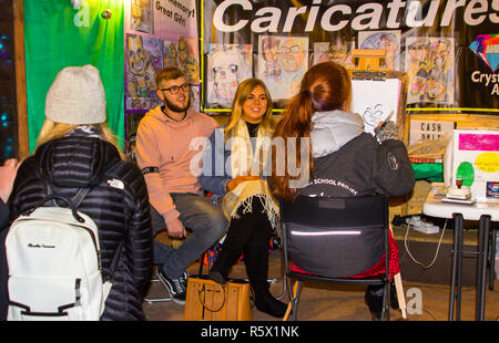 30 November 2018 A young couple sit for a caricature artist in Belfast in a booth at the Belfast City Christmas Fair. The fair is now a popular annual Stock Photo