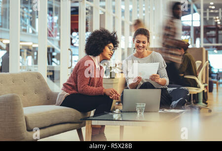 Two businesswomen looking at a paper discussing work at office. Smiling businesswoman sitting with coworker in office with a laptop in front. Stock Photo