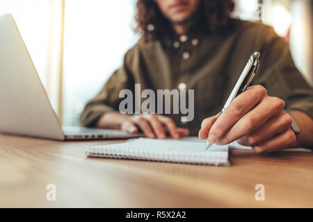 businessman writing in notepad with a laptop on the desk. Close up of hand of businessman holding pen and making notes. Stock Photo