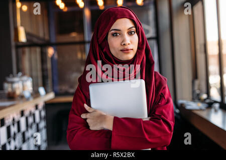 Confident woman in hijab with laptop standing indoors at a restaurant. Islamic businesswoman in hijab at coffee shop. Stock Photo