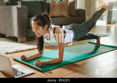 Smiling woman exercising at home and watching training videos on laptop. Chinese female doing planks with a leg outstretched and looking at laptop. Stock Photo