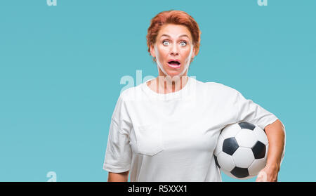 Atrractive senior caucasian redhead woman holding soccer ball over isolated background scared in shock with a surprise face, afraid and excited with f Stock Photo