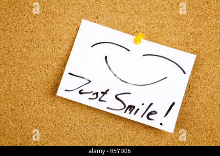 A yellow sticky note writing, caption, inscription Top view of Just Smile sticky note pasted in black ext on a sticky note pinned to a cork notice board Stock Photo