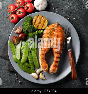 Tasty and healthy salmon steak with green peas, broccoli and tomatoes on a gray plate. Diet food on a dark background. Top view. Flat lay. Stock Photo