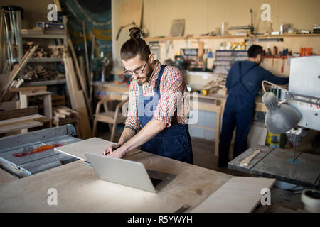 Portrait of two workers in carpenters workshop, focus on modern young man using laptop in foreground, copy space Stock Photo