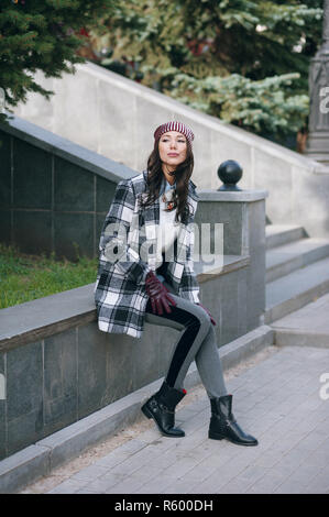 Young beautiful stylish woman, wearing a coat in a white and black cage, white jacket, gray trousers, black boots. Street style, spring and autumn trend, a flirty look Stock Photo