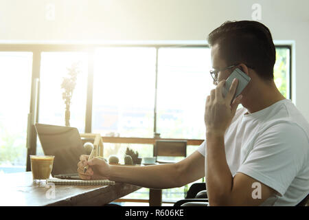 Young asian man talking smartphone and using computer laptop in coffee shop. Stock Photo