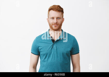 Waist-up shot of attractive masculine macho man with ginger hair in green polo shirt smirking and gazing with confident and self-assured expression at Stock Photo