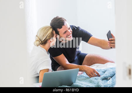 beautiful young millennial caucasian couple play on the bed with technology taking selfie picture with smartphone smiling and having fun together - so Stock Photo