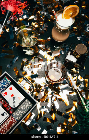 top view of alcoholic cocktails, playing cards, poker chips and party horns on table covered by golden confetti Stock Photo