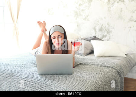 Cheerful brunette woman holding glass of wine wearing sleaping mask in bed. Reading and looking at laptop Stock Photo