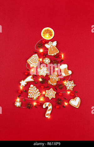 Creative Christmas tree made of iced gingerbread cookies, anise stars, berries, orange chips, decorated red ribbon bow on red background. New Year greeting card concept. Top view. Stock Photo