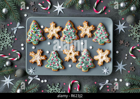 Plate of Xmas cookies shaped as Christmas trees and snowflakes on grey plate. Flat lay, dark background decorated with fir tree twigs, candy canes, st Stock Photo