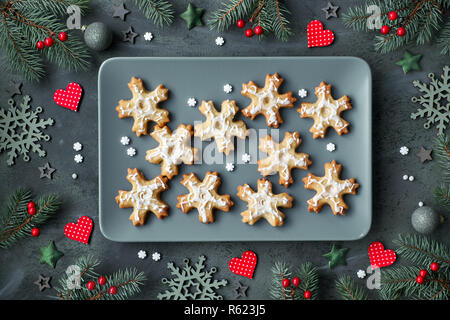 Plate of Christmas cookies shaped as snowflakes on grey plate. Flat lay on dark background decorated with Xmas tree twigs, hearts, berries and stars. Stock Photo