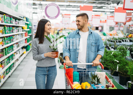 Couple with cart full of goods buying home flower in a supermarket, family shopping. Customers in shop, buyers in market Stock Photo