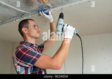 Young man in goggles fixing drywall suspended ceiling to metal frame using electrical screwdriver on ceiling insulated with shiny aluminum foil. Renov Stock Photo
