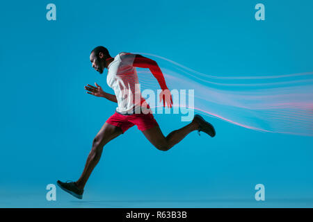 Full length portrait of active young african muscular running man, isolated over blue studio background with flashes of light Stock Photo