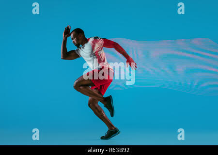 Full length portrait of active young african muscular running man, isolated over blue studio background with flashes of light Stock Photo