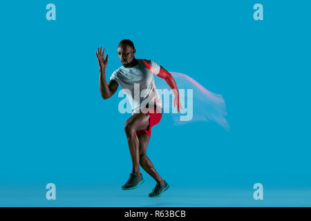 Full length portrait of active young african muscular running man, isolated over blue studio background with flashes of light Stock Photo