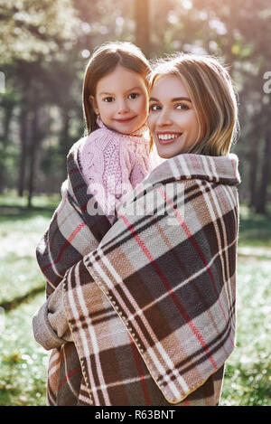 A little curly girl in sweater is hugging her mother in woods. She is wearing pink sweater and young woman is in grey sweater. They are wrapped up wit Stock Photo