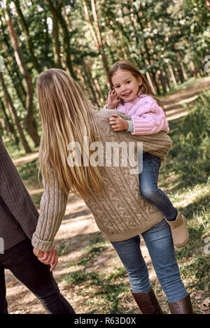 Mother holds laughing daughter on hands in forest near father. Stock Photo