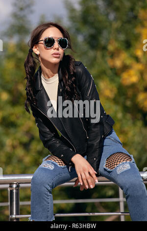 fashionable young woman in rock style clothes, black leather jacket, blue jeans, tights in a grid under battered jeans Stock Photo