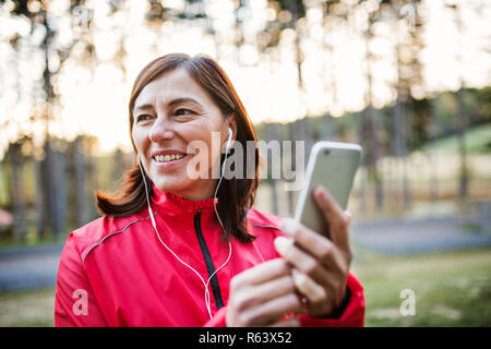 A female runner with earphones outdoors in autumn nature, using smartphone. Stock Photo