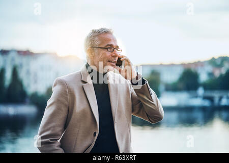 Mature businessman with smartphone standing by river at sunset, making phone call. Stock Photo