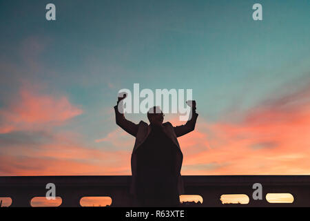 A silhouette of businessman standing on a bridge at dusk, expressing excitement. Stock Photo