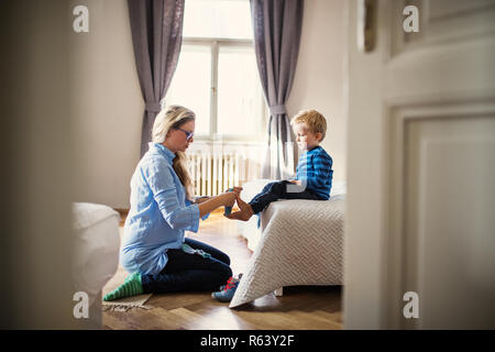 A young mother putting socks on toddler son inside in a bedroom. Stock Photo