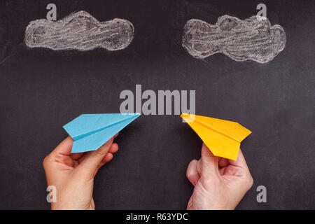 Two hands holding two colorful paper planes under clouds. One paper plane is blue, the other is yellow. Stock Photo