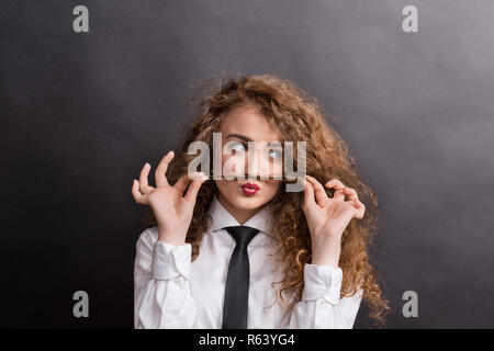 Young woman in studio, wearing white shirt and black tie, making fake mustaches. Stock Photo