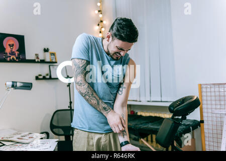 Confused dark-haired man aggressively scratching his primitive tattoo Stock Photo